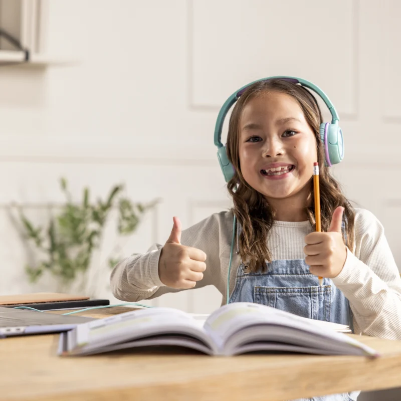 A smiling young girl wearing headphones sits at a table with an open book and a laptop, giving two thumbs up. She appears to be studying on an online learning platform in a bright, cozy room.