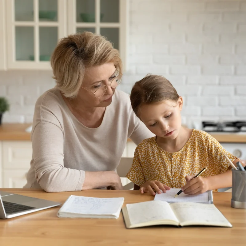 An older woman and a young girl sit at a kitchen table. The woman watches as the girl writes in a notebook, creating an atmosphere reminiscent of a Learning Hub. A laptop, papers, and a pencil holder are on the table, with the kitchen in the background.