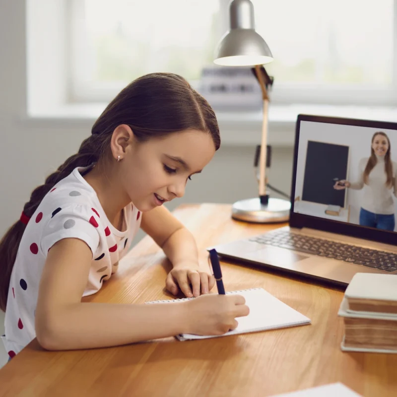A young girl sits at a desk, writing in a notebook while attending an online class on her laptop. A woman appears on the screen, teaching and demonstrating how Learning Hub works, with books and a green apple on the desk.