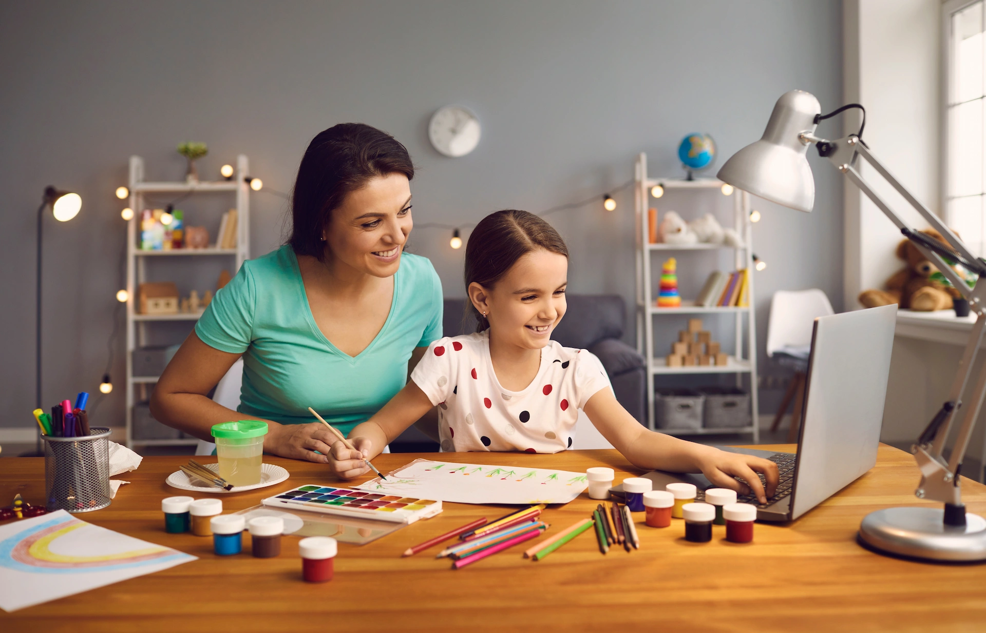 A woman and a young girl sit at a desk doing arts and crafts with paints and colored pencils, smiling and looking at a laptop—perhaps reviewing measures of academic progress—in a cozy, well-lit room with shelves and art supplies in the background.