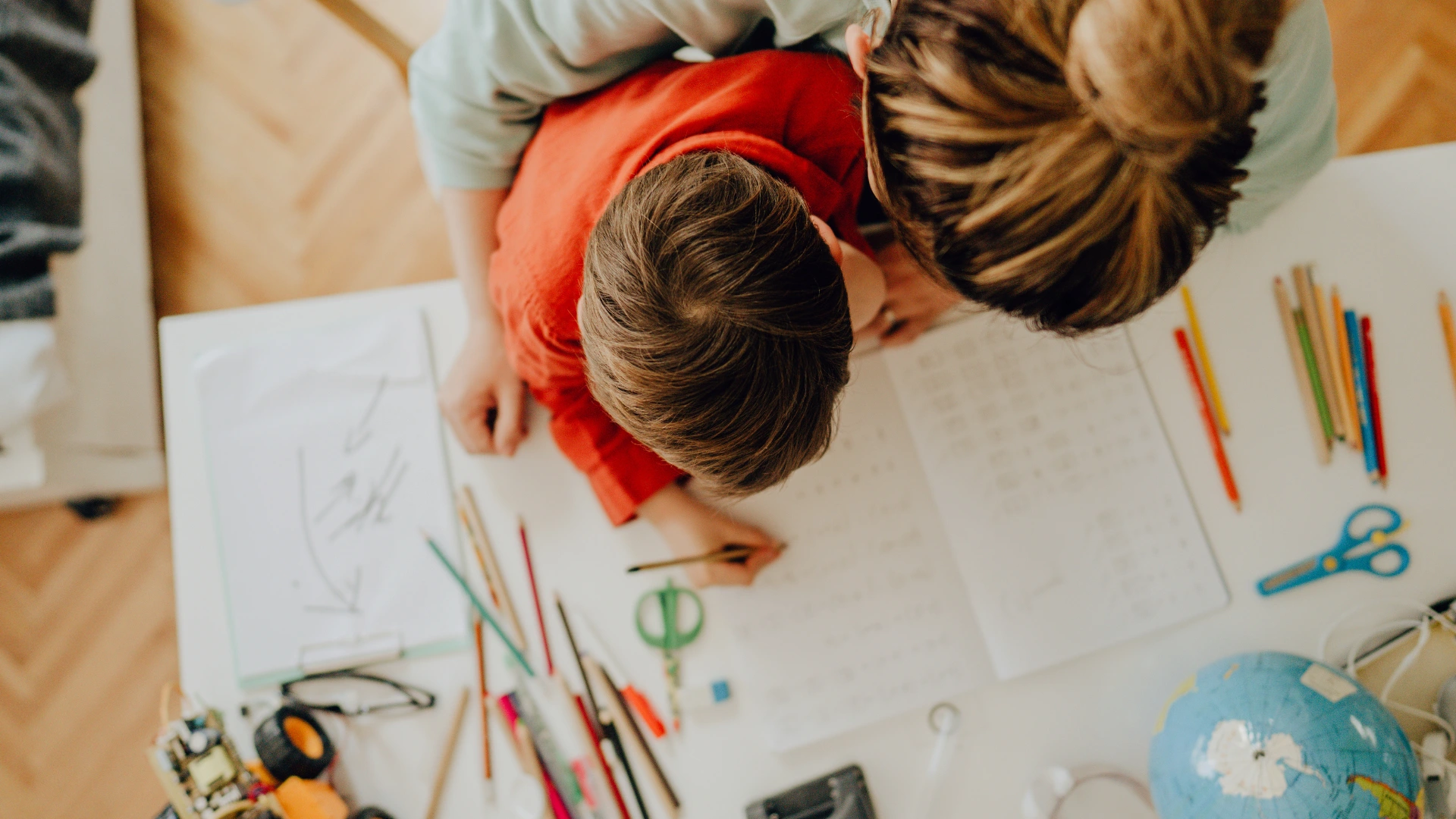 An adult helps a child with homework at a white desk covered in papers, colored pencils, and school supplies, viewed from above. Find Your Familys Flow as the child writes in a workbook while the adult looks on.