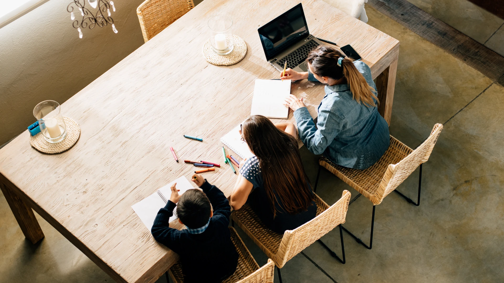 Overhead view of a woman and two children sitting at a wooden table, writing or drawing on paper with colored pens. A laptop and cup are also on the table, reflecting a bright, homey atmosphere within a close-knit homeschool community.