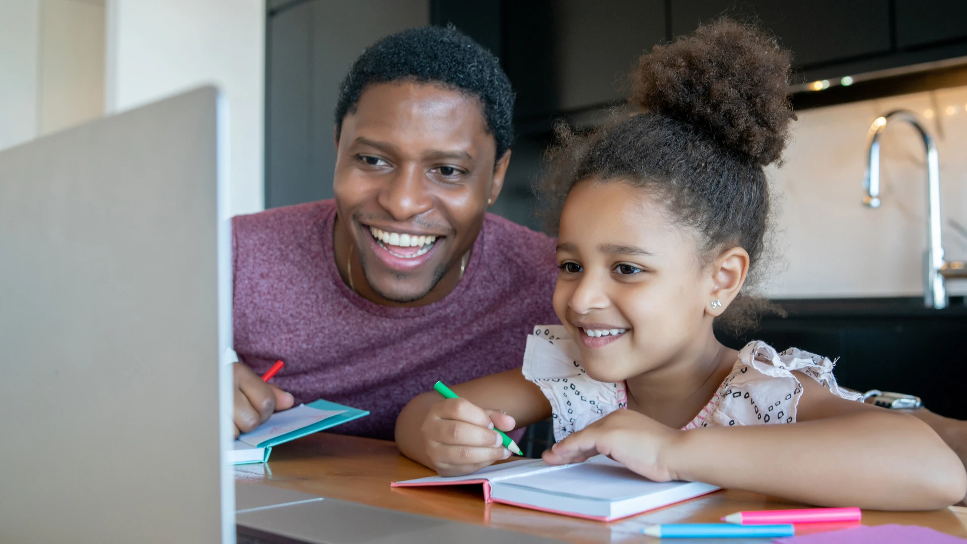 A man and a young girl smile while sitting at a table, writing in notebooks and looking at a computer screen, highlighting the Homeschool Co-op Benefits of collaborative online learning.