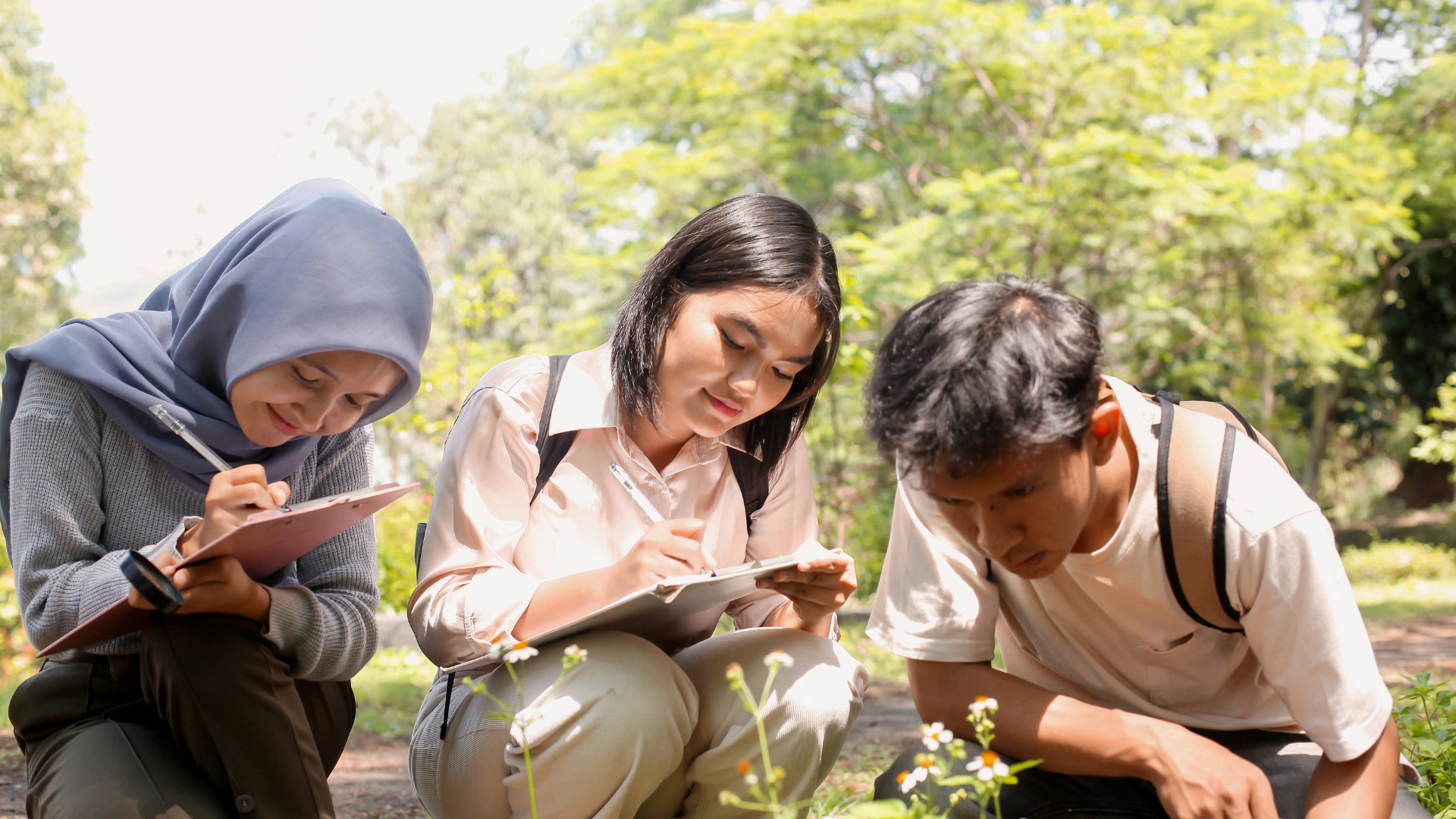 Three young people sitting outdoors, closely observing and writing notes on clipboards while studying plants; they appear to be sharing Nature Exploration Tips amid trees and bright sunlight in the background.