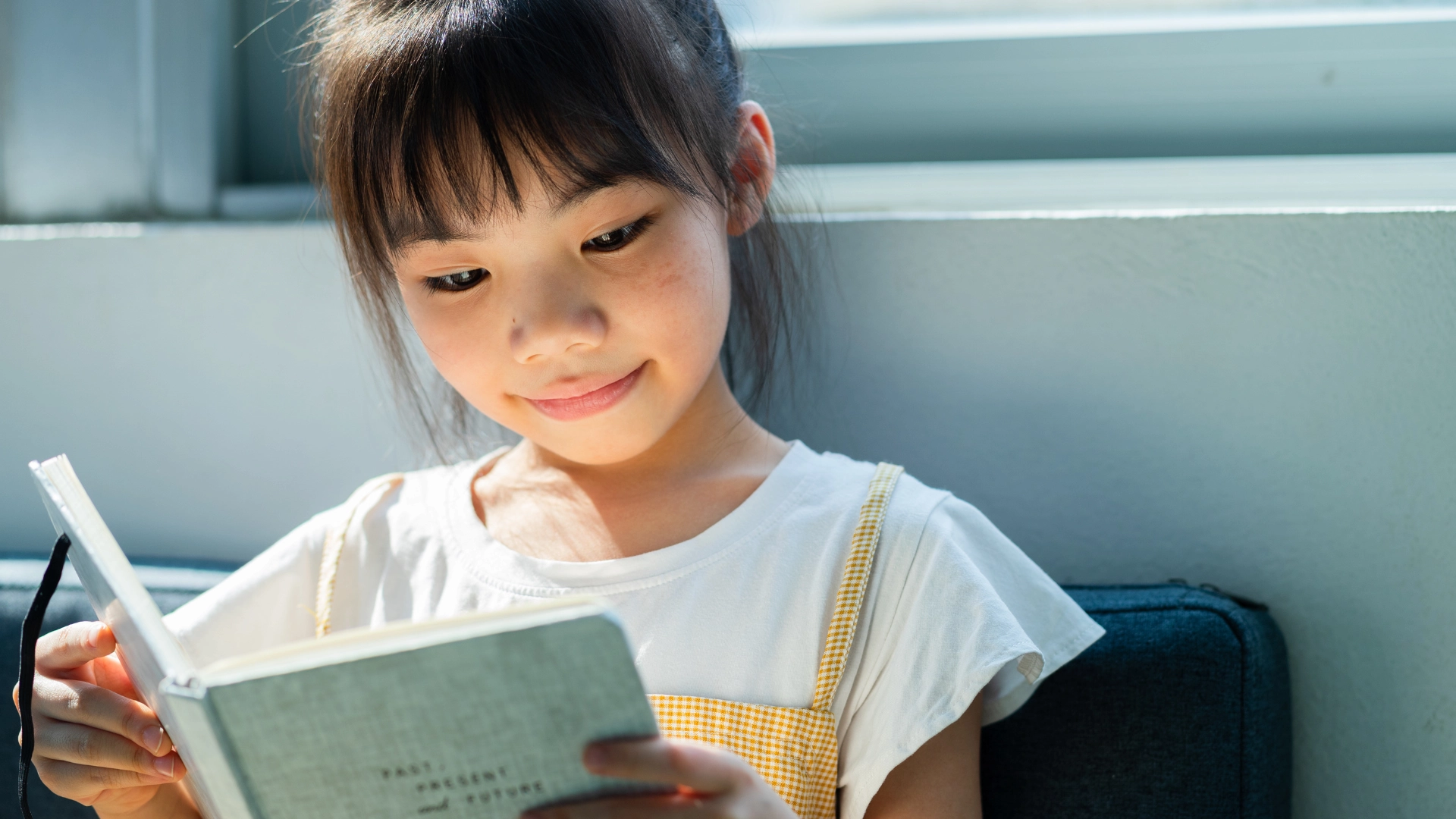 A young girl with dark hair and bangs, wearing a white shirt and yellow suspenders, sits indoors by a window and smiles while reading a book. Sunlight shines on her face.
