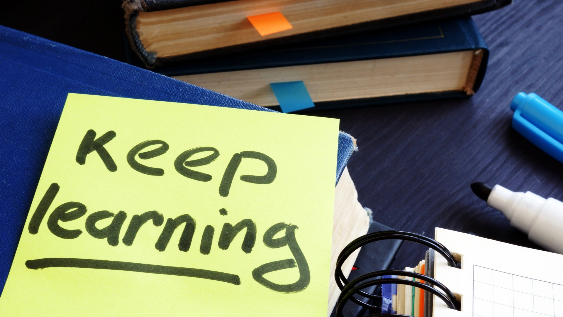A sticky note with the handwritten message Keep learning sits on top of a book, surrounded by stacked books, colorful bookmarks, a spiral notebook, and a marker—perfect for inspiring any homeschool learning style.