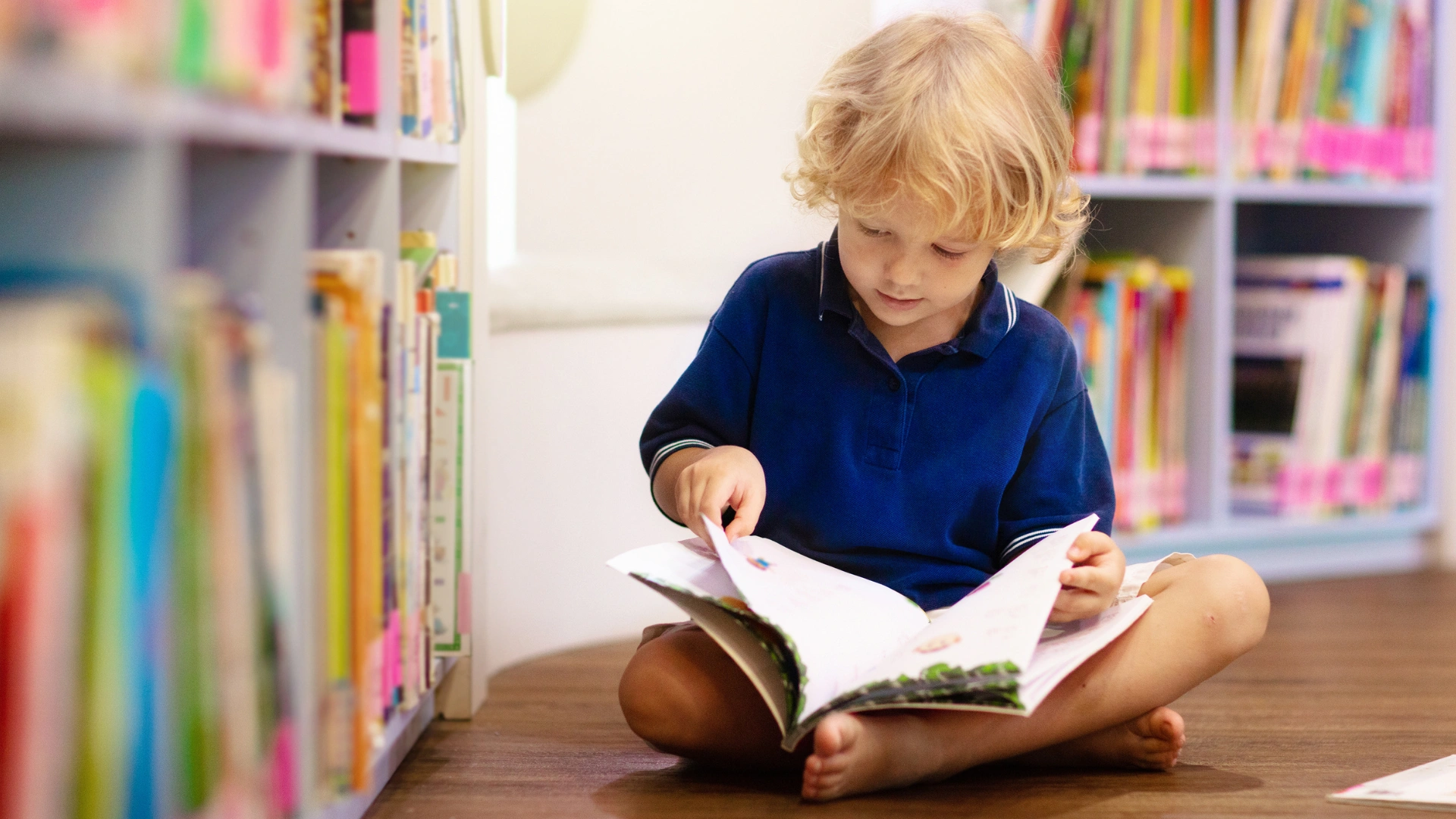 Young child with blonde hair, wearing a blue shirt, sits cross-legged on a wooden floor in a library, exploring an open book—an ideal setting for a Homeschool Reading Curriculum—surrounded by bookshelves filled with colorful books.