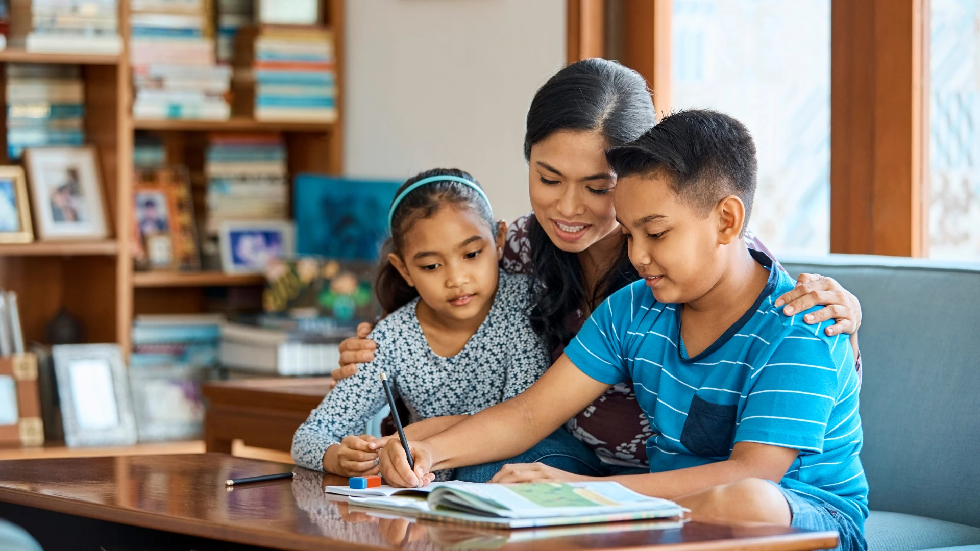 A woman sits at a table with two children, helping them with homework. One child writes in a notebook while the other looks on. Bookshelves are visible in the background, and they all appear focused and engaged.