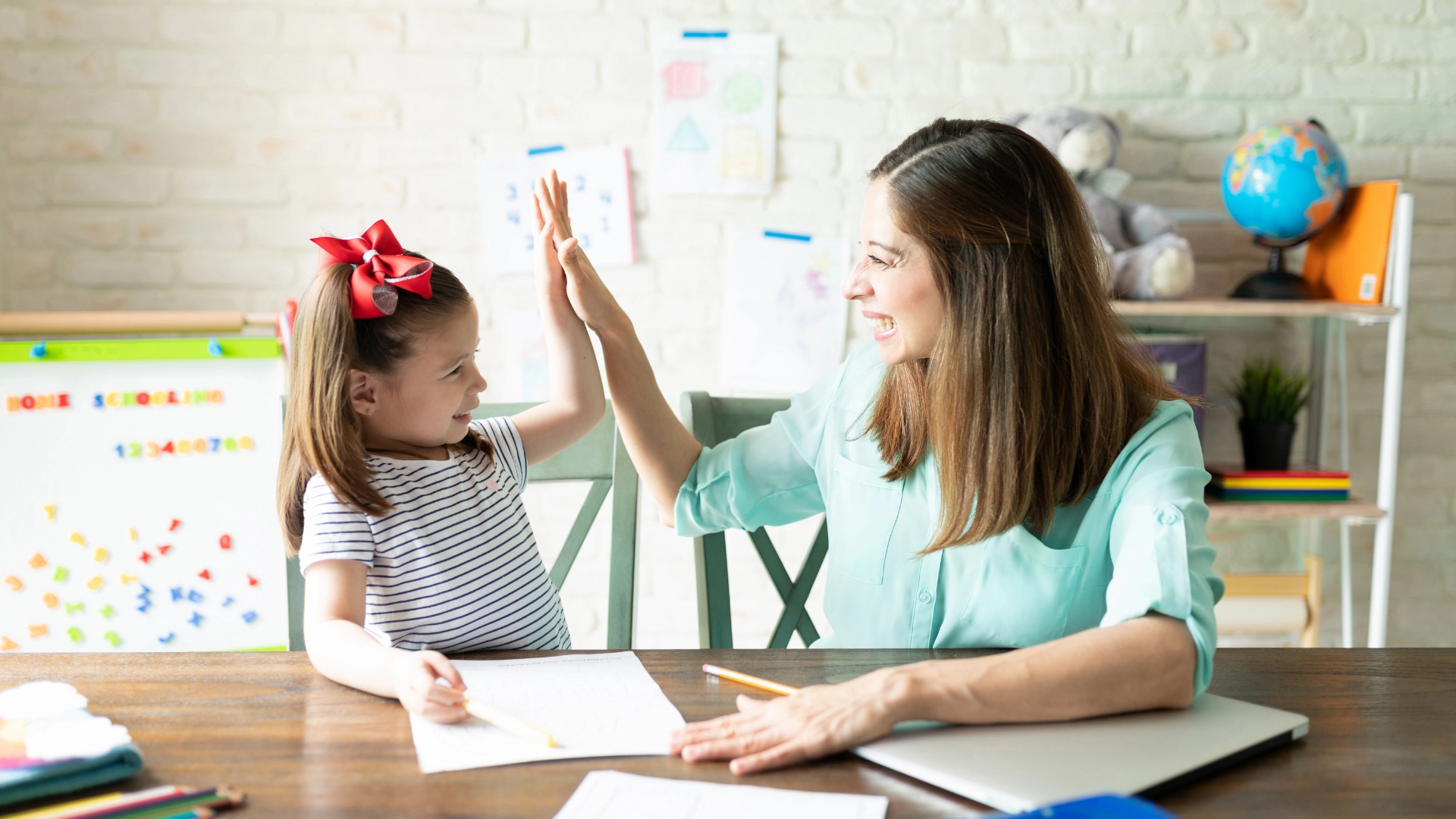 A young girl and an adult woman sit at a table, smiling and giving each other a high-five while using Homeschool Resources for homework or drawing, with school supplies and colorful artwork in the background.