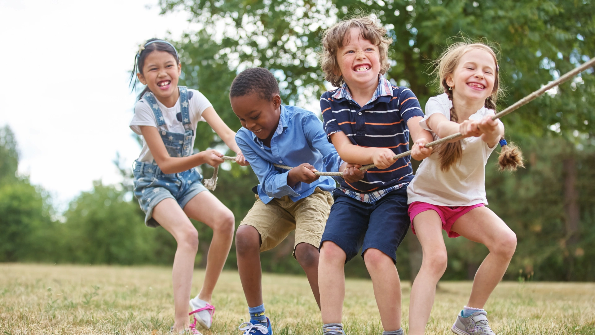 Four children laughing and smiling while playing tug-of-war outdoors on a grassy field, pulling a rope together on a sunny day with trees in the background.