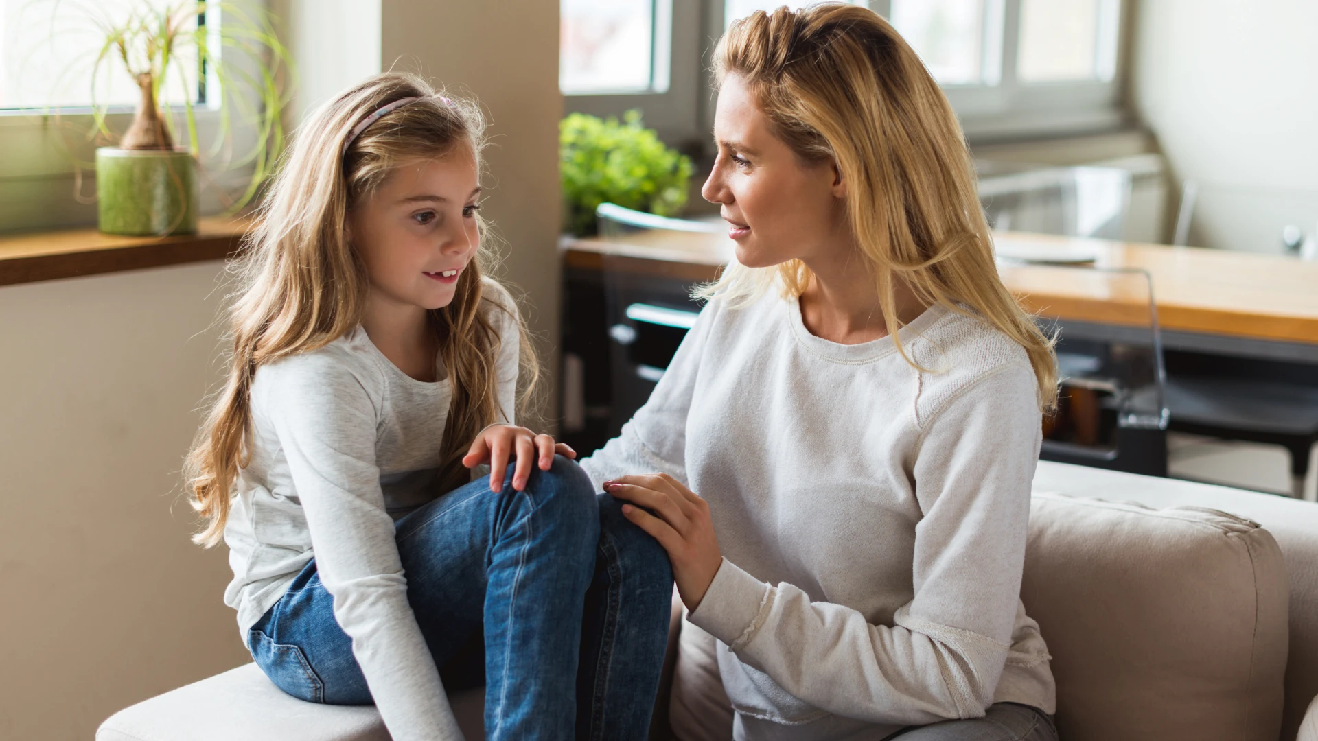 A woman and a young girl sit on a couch indoors, facing each other and having a conversation to improve reading fluency. Both have long blonde hair and are wearing light-colored tops and jeans as sunlight streams in through the windows behind them.