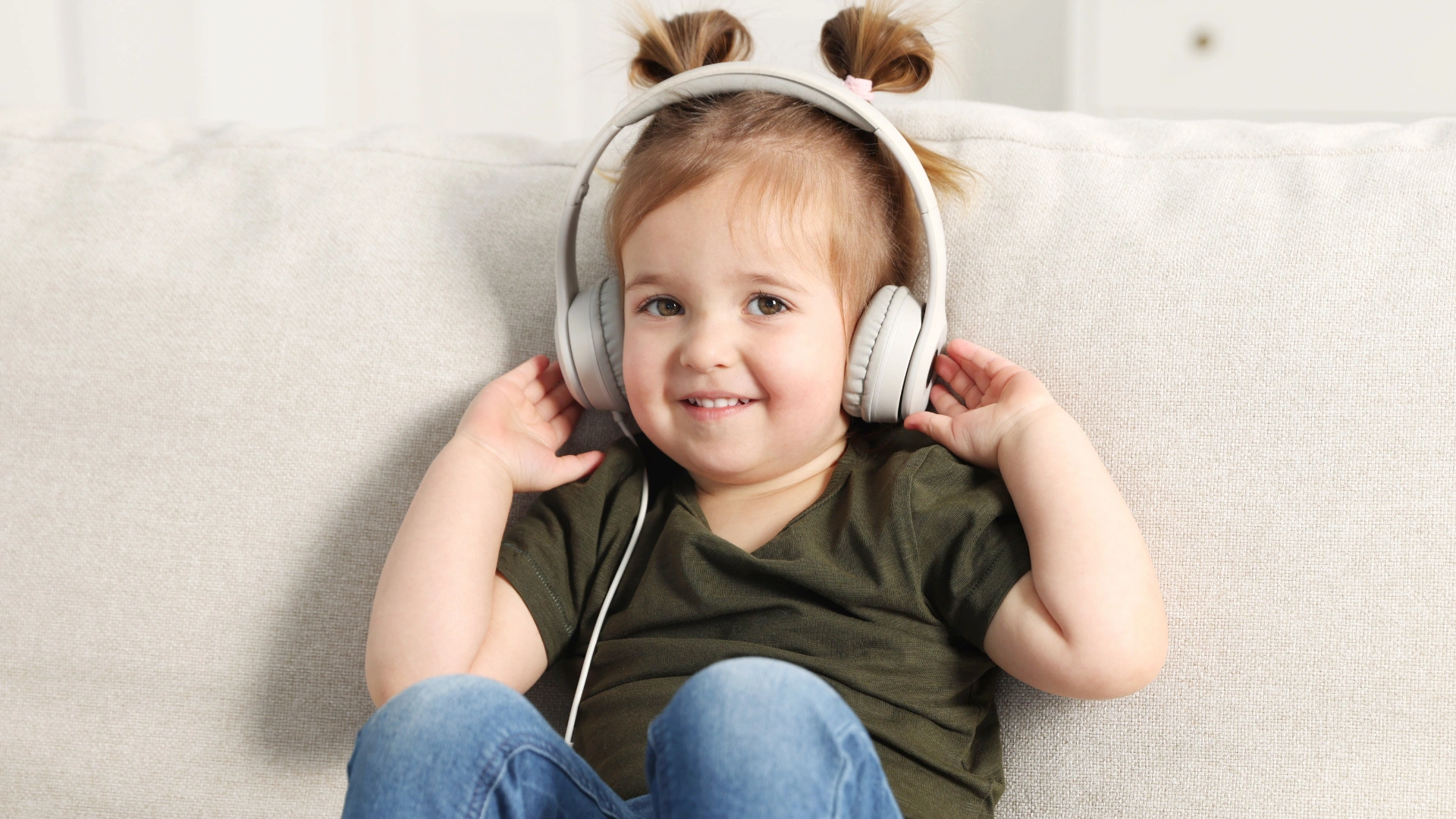 Young child with light brown hair in two pigtails sits on a light-colored couch, wearing a dark green shirt and blue jeans, smiling and listening to audiobooks with large white headphones for engaging reading comprehension activities.