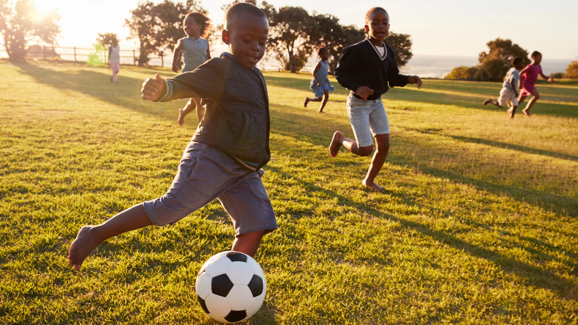 A group of children play soccer on a grassy field at sunset, enjoying one of the best extracurricular ideas for homeschoolers. One child in the foreground prepares to kick the ball, while others run happily amid trees and golden sunlight.
