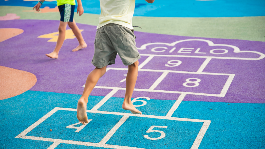 Two barefoot children play hopscotch on a colorful playground, jumping on numbered squares. The word CIELO is written inside a cloud shape at the end of the hopscotch path.
