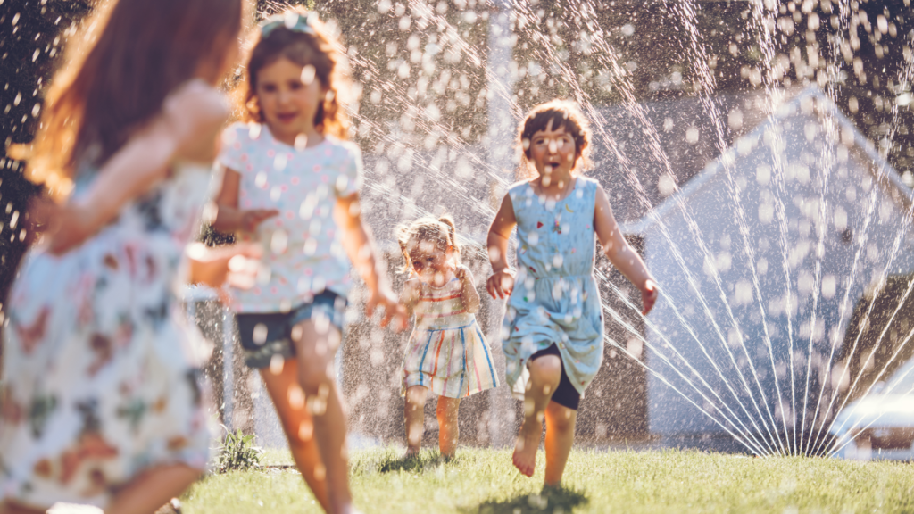 Four young children joyfully run through a backyard sprinkler on a sunny day, with water droplets sparkling in the sunlight and the grass beneath their feet.
