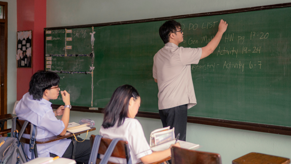 A teacher writes a DO LIST on a chalkboard in a classroom while two students in uniform sit at desks, reading and taking notes. The classroom has a green chalkboard and educational materials.